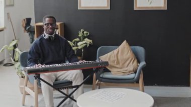 Gündüz vakti modern apartman dairesinde sentezleyicinin önünde oturan, kameraya poz veren ve gülümseyen gözlüklü ve kulaklık takan yakışıklı siyahi bir adamın orta boy portresi.