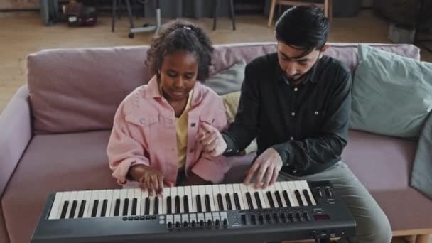 高角のプロフェッショナル男性アジアの音楽教師助けて黒10歳の女の子遊びますシンセサイザー上のソファに座って自宅で午後 — ストック動画
