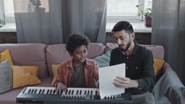 Profesyonel Asyalı erkek akıl hocası meraklı küçük zenci çocuğa müzik notalarını öğretiyor. Gündüz vakti apartmanda sentezleyicinin önünde oturuyor.