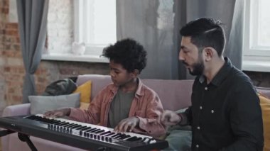 Profesyonel Asyalı erkek öğretmenle sentezleme eğitimi alan kararlı küçük zenci çocuk öğleden sonra evde kanepede oturuyor.