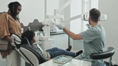 Orta uzunlukta, ofisinde önlük, eldiven ve yüz maskesi takmış, dişçi koltuğunda oturan siyah ilköğretim çocuğunda ağız aynası kullanan, annesinin yanında durduğu beyaz bir erkek doktor.