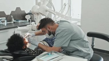 Önlük, maske ve eldiven takmış, dişçi koltuğunda uzanan siyah çocuğu tedavi eden orta boylu beyaz erkek doktor.