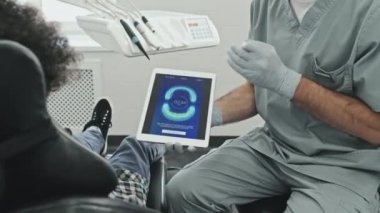 Ameliyat önlüğü ve eldiven giyen orta boylu erkek doktor tablet bilgisayarın ekranında gündüzleri dişçi koltuğunda oturan kıvırcık saçlı hastaya mükemmel ısırık görüntüsü veriyor.