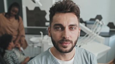 Sakallı beyaz erkek dişçinin yakın plan portresi. Önlük takıyor, kameraya gülümsüyor ve parlak ofisinde poz veriyor. Arka planda siyah kadın ve oğlunu bulanıklaştırıyor.