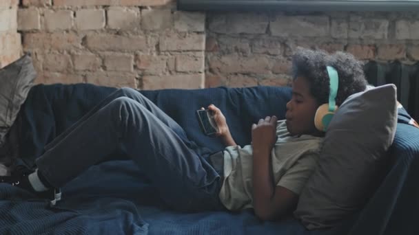 10岁的黑人男孩头戴耳机 白天躺在房间沙发上 有棒棒糖 使用智能手机 全景全景 — 图库视频影像