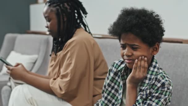 黑人10岁男孩牙疼 坐在诊所候诊室里 与使用智能手机的年轻妈妈交谈 进行胸部检查 — 图库视频影像