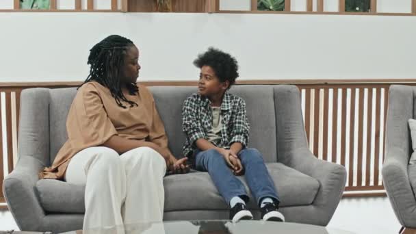中等身材的年轻黑人妇女和她十岁的儿子坐在接待室的沙发上聊天 — 图库视频影像