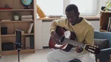 Orta uzunlukta siyah bir adam gündüz vakti oturma odasında oturuyor, akustik gitar çalıyor, akıllı telefon kamerasıyla konuşuyor, canlı yayın yapıyor.