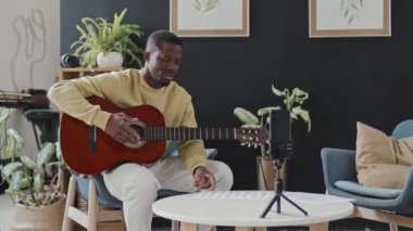 Orta uzunluktaki genç siyahi adam gündüz vakti koltukta oturuyor, akustik gitar çalıyor, gülümsüyor ve akıllı telefon kamerasıyla konuşuyor, çevrimiçi video yayınlıyor.