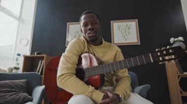 Gündüz vakti modern bir dairede oturan, akustik gitar çalan, kameraya bakıp konuşan neşeli siyahi adamın düşük açılı portresi.