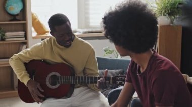 Gündüz vakti oturma odasında oturan kıvırcık saçlı genç adamın akustik gitarda akor gösteren ve konuşan siyah erkek müzik öğretmenine bakışının yan görüntüsü.