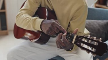 Gündüz vakti evdeki koltukta oturup akustik gitar çalan kırpık bir adamın orta boy yakın çekimi.