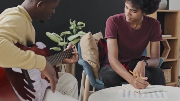 집에서 의자에 교사가 어쿠스틱 기타를 연주하고 이야기할 필기를 곱슬머리의 청년의 — 비디오