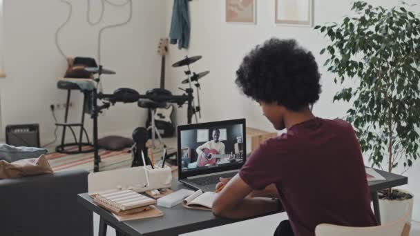 ギターを弾いている黒人男性とラップトップコンピュータ上のオンライン音楽のレッスンを持っているときにノートを取り 午前中に彼の部屋に机の上に座っている巻き毛の若い男の背面図 — ストック動画