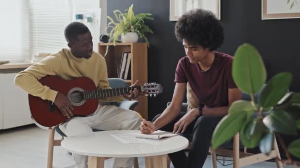 中等身材 卷曲头发的年轻人坐在客厅的扶手椅上 在黑人音乐老师用声吉他弹奏和弦的时候做笔记 — 图库视频影像