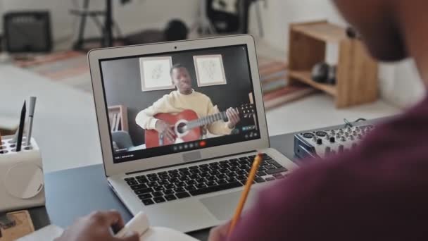在他的房间里 坐在书桌前的一个剪贴画的学生的肩膀上 当视频通过手提电脑与会弹吉他的黑人通话时 他们会做笔记 — 图库视频影像