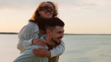 Yüzünde sakalı olan mutlu genç bir adam neşeli kız arkadaşını sırtında taşıyor doğada buluşmanın tadını çıkarıyor.