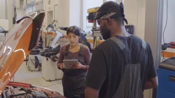 西班牙裔女机修工身穿制服 使用数字平板电脑 与非洲裔美国男性同事讨论汽车故障问题 同时在维修车库一起工作 — 图库视频影像