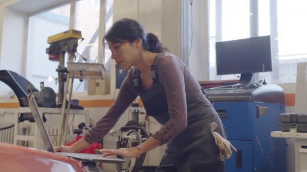 穿着制服的西班牙女人一边在汽车修理店当机械师 一边用笔记本电脑做汽车诊断 — 图库视频影像