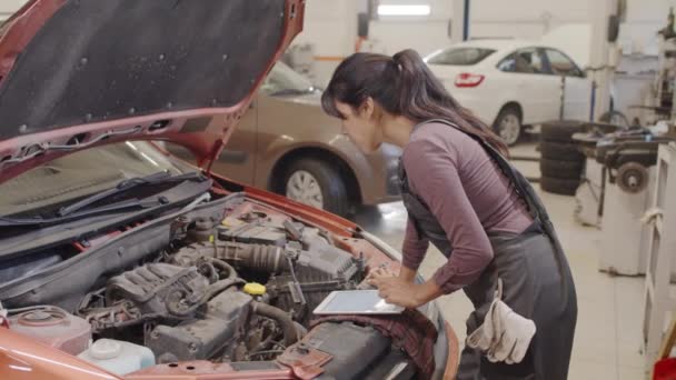 Kobieta Mechanik Badania Części Samochodowych Pod Maską Przy Użyciu Tabletu — Wideo stockowe