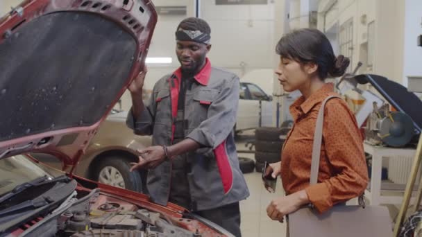 非裔美国人汽车修理工在车篷下展示汽车零部件 并向维修车库的女客户解释故障原因 — 图库视频影像