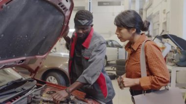 Araba parçalarını kaput altında gösteren Afrikalı Amerikalı tamirci ve oto servis garajındaki İspanyol bayan müşteriye sorunları açıklıyor