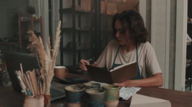 Önlük giyen, çömlekçilik atölyesinde masaya oturan, not alan ve gündüz bilgisayar kullanan beyaz bir kadının sandığı.