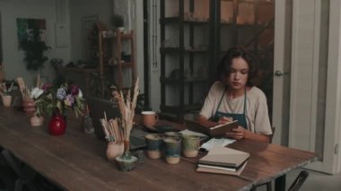 Önlük giyen, çömlekçilik atölyesinde masaya oturan, not defterine not alan ve gündüzleri bilgisayar kullanan beyaz kadın girişimcinin sandığı.