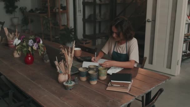 白种人年轻的女经理穿着围裙 坐在陶器作坊的桌旁 在笔记本里记笔记 白天用电脑 — 图库视频影像