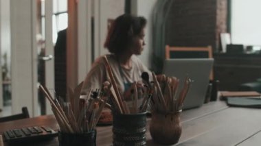 Gün içinde atölyedeki üç kavanozda birden fazla boya fırçasının seçici odak noktası, arka planda oturan ve çalışan bulanık kadın.