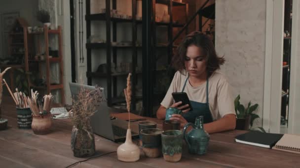 若い女性のウエストアップスローモーションテーブルに座っている白人の陶器店の労働者 オンラインショップのための手作りの陶器の写真を撮る スマートフォンを使用して — ストック動画