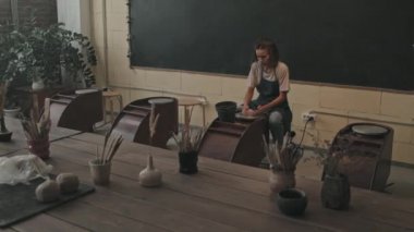 Önlük ve tişört giyen kahverengi saçlı genç bir kadının modern atölyede oturup çömlek çarkında kil şekillendirdiği geniş bir kare.