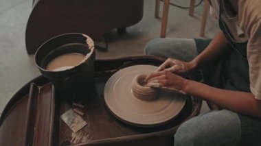 Önlük ve tişört giyen, modern atölyede oturan, çömlek çarkında kil şekillendiren, seramik yapan...