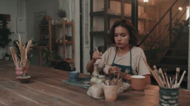 Yüzünde kuru kil olan, önlük takan, modern stüdyoda masada oturan, fırçayla el yapımı seramik vazo boyayan, dikkatli beyaz kadın sandığı.