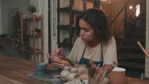 穿着围裙 头戴干粘土 细心的棕色头发高加索女人 坐在现代工作室的桌子旁 用刷子画手工制作的陶瓷花瓶的胸部慢动作 — 图库视频影像