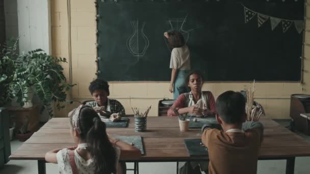 四个不同的孩子 中等个子 坐在陶艺学校的桌旁 揉碎生土 塑制陶瓷制品 白种人女老师在黑板上画花瓶 — 图库视频影像
