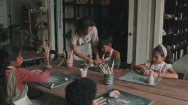 Orta uzunlukta beyaz kadın seramikleri çömlekçilik okulunda masada oturan, çiğ kil parçalarını yoğuran, gündüz seramik öğeleri şekillendiren küçük çocuklara yardım ediyor.