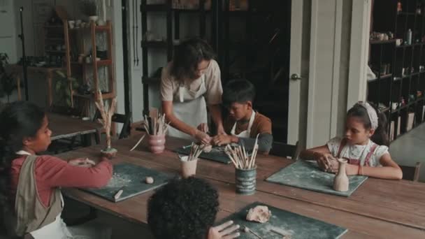 中等长度的白人女神学家帮助在陶器学校坐在桌旁 揉碎生土 白天塑制陶瓷制品的幼儿 — 图库视频影像