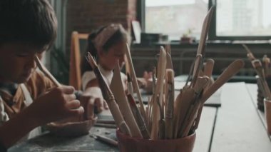 Asyalı bir erkek ve beyaz bir kızın çömlekçilik okulunda masada oturup, tahta aletlerle kil şekillendirmesi, gündüz seramik eşyalar yapması, çömlek yapmayı öğrenmesi,