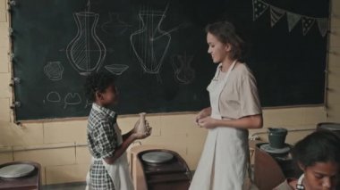 Kareli gömlek ve önlük giyen küçük siyahi çocuğun izini sürüp kilden yapılmış vazosunu sınıftaki tahtası önünde duran beyaz kadın öğretmene gösteriyordu.