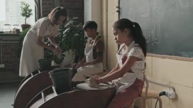 Orta uzunlukta önlük takan, çömlek tekerlerinde oturan, kilden el sanatları yapan, gündüz vakti gezen ve onlara öğretmenlik yapan çocuklar.