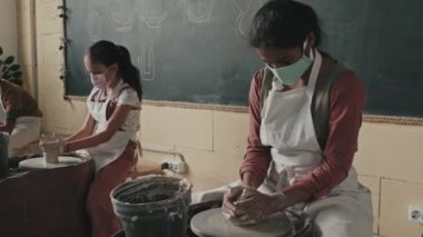 Orta uzunluktaki Afrika kökenli Amerikalı kız önlük ve yüz maskesi takıyor, gündüz atölyesinde çömlekçi çarkında oturup el işi yapıyor.