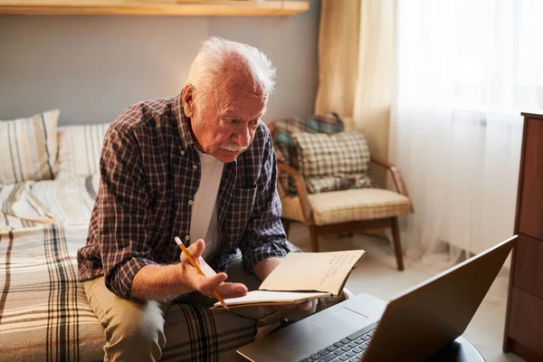 Hombre mayor con cuaderno y lápiz sentado frente a la computadora portátil — Foto de Stock