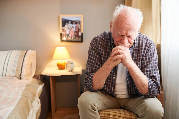 Ηλικιωμένος άνδρας με καθημερινά ρούχα κάθεται στην πολυθρόνα στην κρεβατοκάμαρά του και προσεύχεται — Φωτογραφία Αρχείου