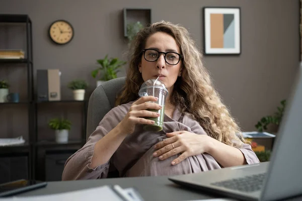 Hamile iş kadını dizüstü bilgisayarın önünde oturmuş smoothie içiyor. — Stok fotoğraf