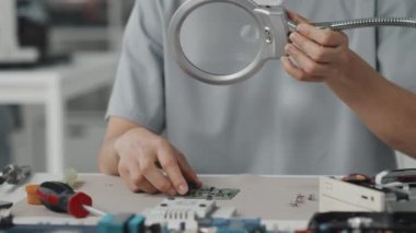 Kadın laboratuvar teknisyeninin masada oturup büyüteçle devre kartını incelerken yakın plan çekimleri.