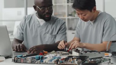 Asyalı kadın mühendisin multimetreyi test çipi olarak kullanırken ve laboratuvarda dizüstü bilgisayar kullanırken Afrikalı Amerikalı meslektaşıyla konuşurken görüntüyü kaldır.