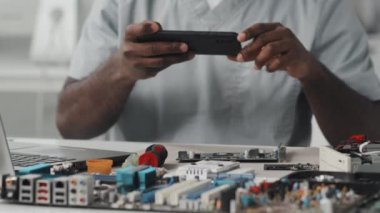Afrikalı Amerikalı teknisyenin, laboratuvarda çalışırken akıllı telefonuyla elektronik bileşenlerin fotoğrafını çekerken görüntüyü yukarı kaldır.