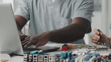 Laboratuvarda yanında oturan ve elektronik aletleri çoklu metre ile inceleyen kadın mühendis bilgisayarında yazı yazan Afrikalı Amerikalı adamın el görüntülerini yakından çek.