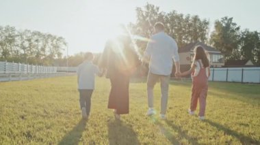 Anne, baba, küçük kız ve oğuldan oluşan beyaz bir ailenin arka planda el ele tutuşması ve güneşli bir günde bahçedeki çimlerin üzerinde yürümesi.